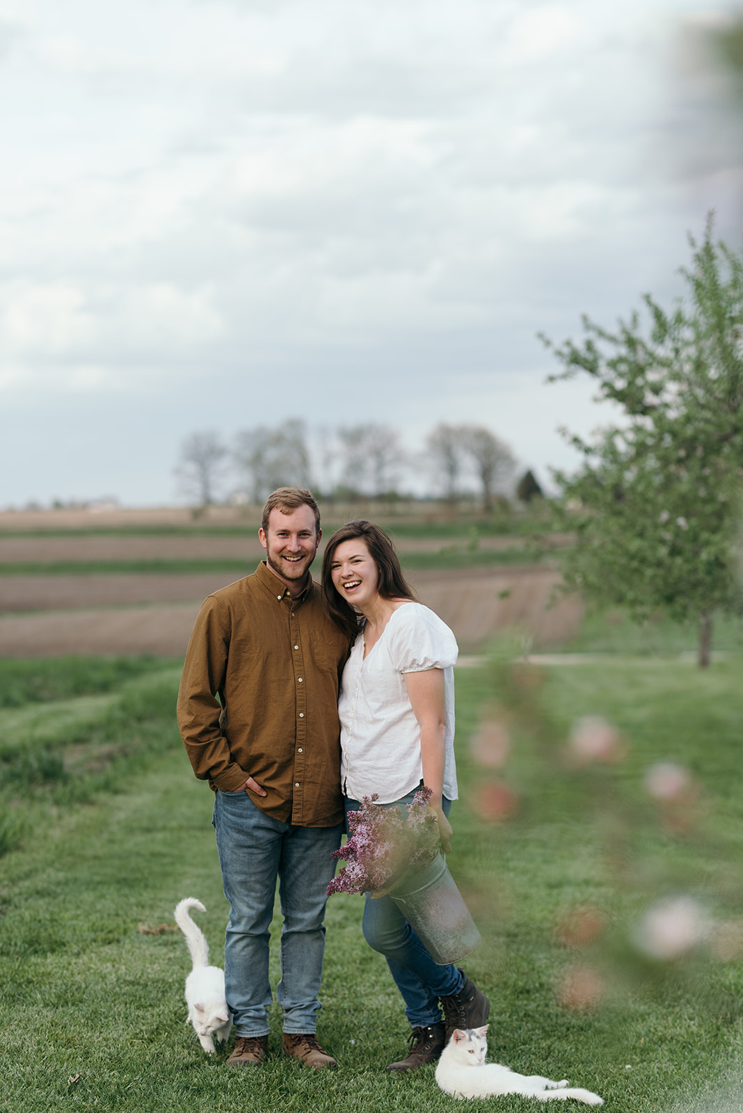 couple-standing-in-field-holding-bucket-of-lilacs-with-cats-wellman-iowa-engagement-session
