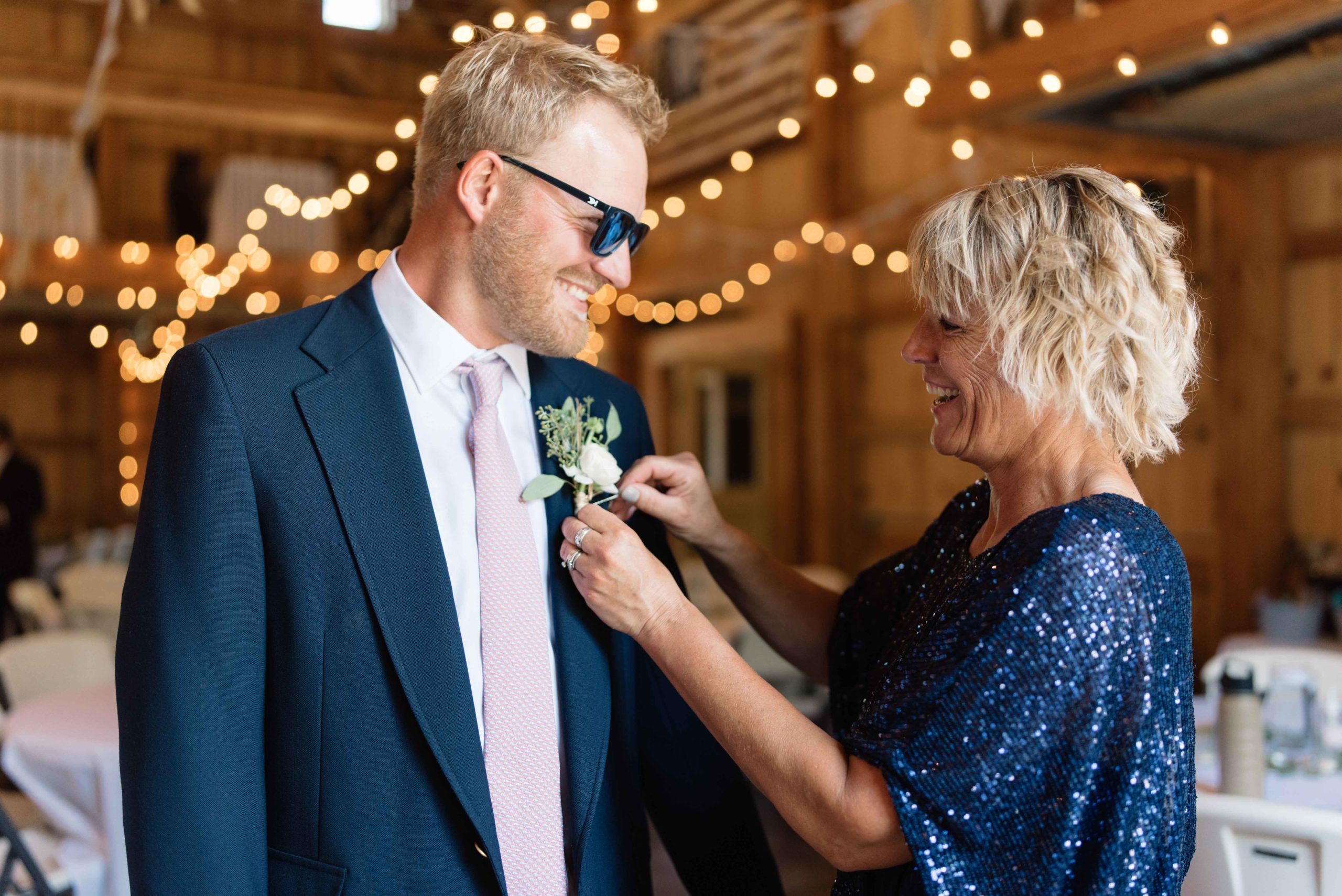 mom putting on grooms boutonniere with edison bulb lights schafer century barn Iowa wedding venue